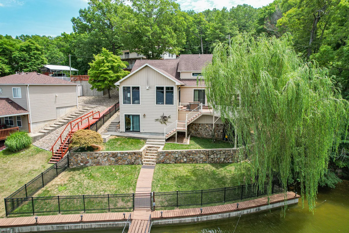 A broad view of the waterfront property showing a spacious deck shaded by a weeping willow, a stone wall, and a staircase leading down to the water. The scene is tranquil and ideal for morning coffee, fishing, or simply enjoying the view.