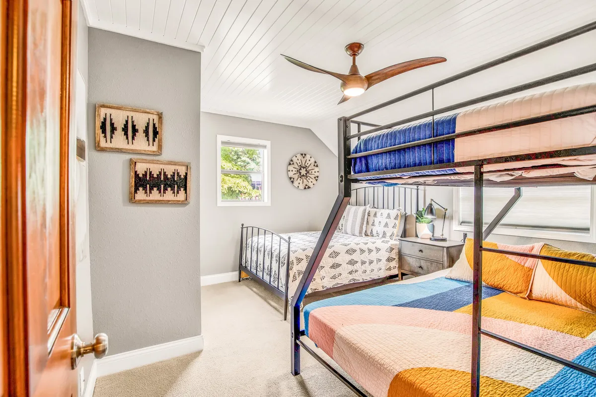 A playful family bedroom featuring a bunk bed and a bed with a patterned quilt, cheerful wall art, and a light, airy atmosphere. The room feels inviting for both children and families, offering versatility for different age groups.
