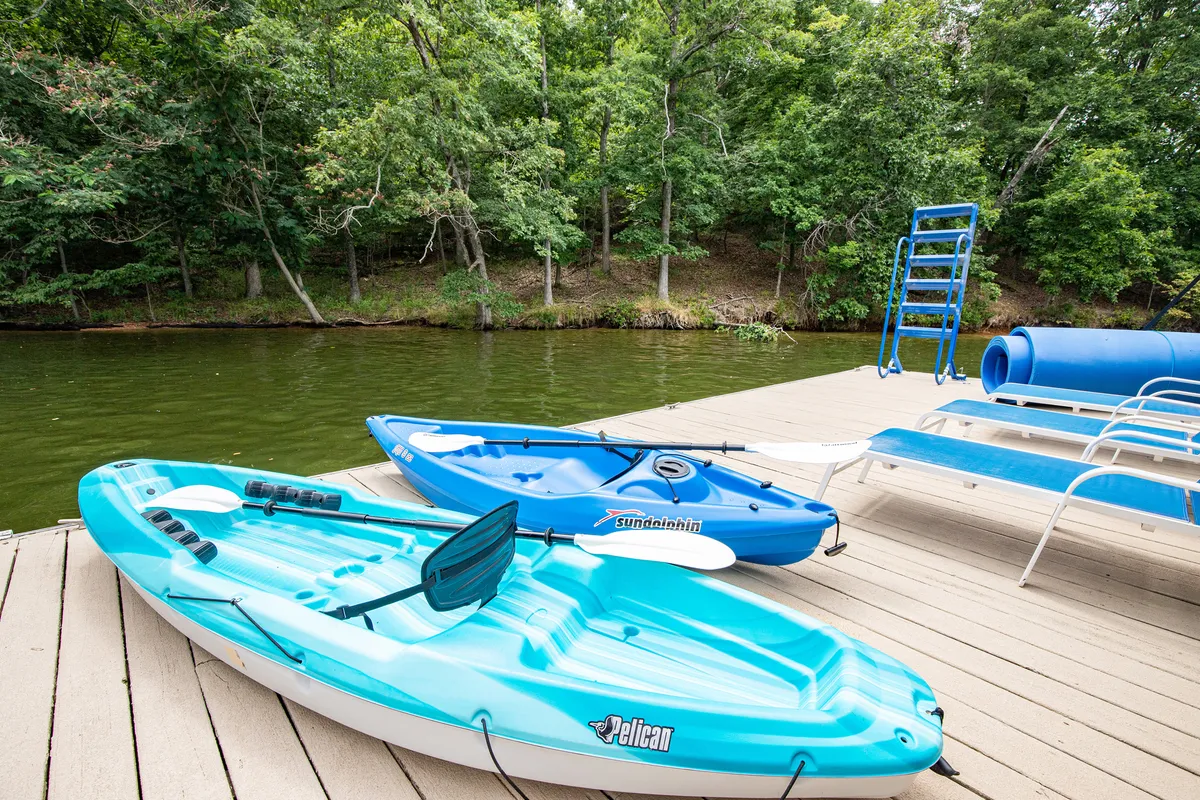 This image captures a vibrant lakefront dock scene featuring two colorful kayaks, a dive platform, and inviting lounge chairs. The setting is bright and peaceful, with calm water in the background ideal for swimming, kayaking, or simply relaxing on the dock.
