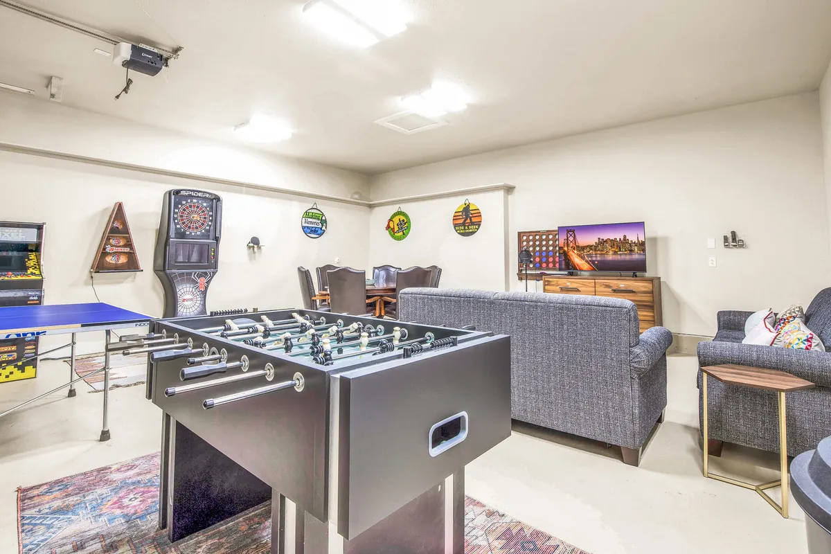 A lively game room featuring a foosball table, dartboard, television, seating area, and a ping pong table. The arrangement suggests a space designed for active play and socializing, with comfortable seating for relaxing or watching TV between games.