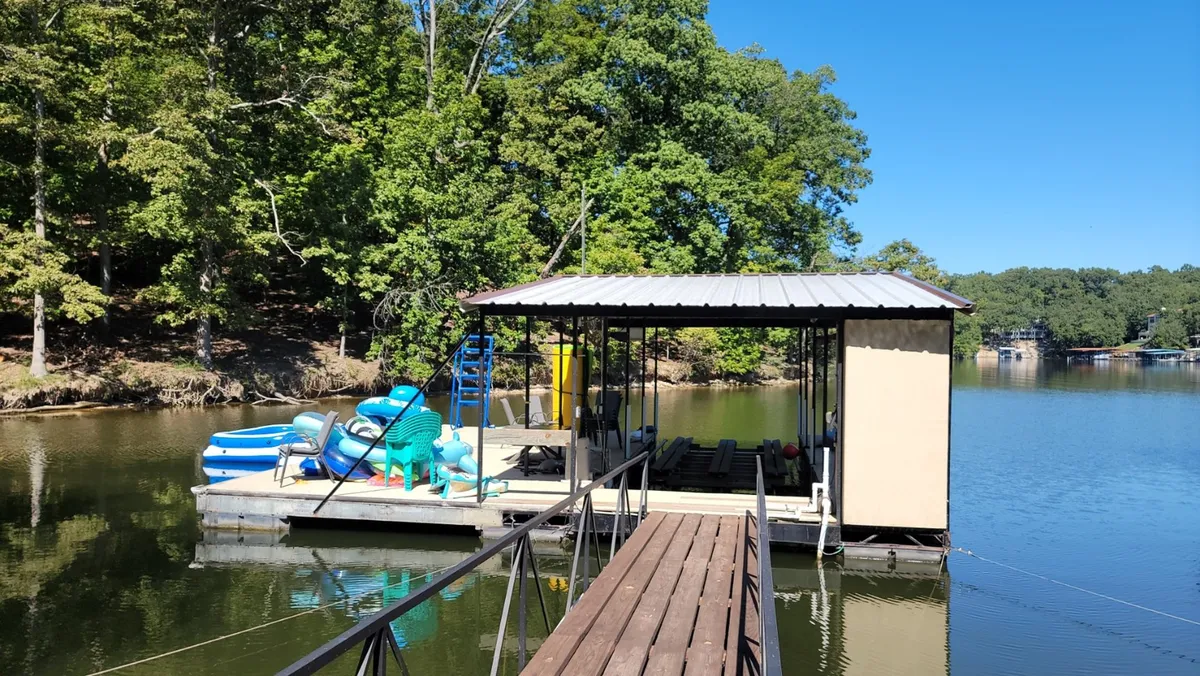A peaceful lakeside dock with a boathouse and kayaks, surrounded by mature trees. The dock extends into the lake, inviting activities like boating, fishing, and relaxing by the water, with a tranquil mood that speaks to leisurely afternoons.