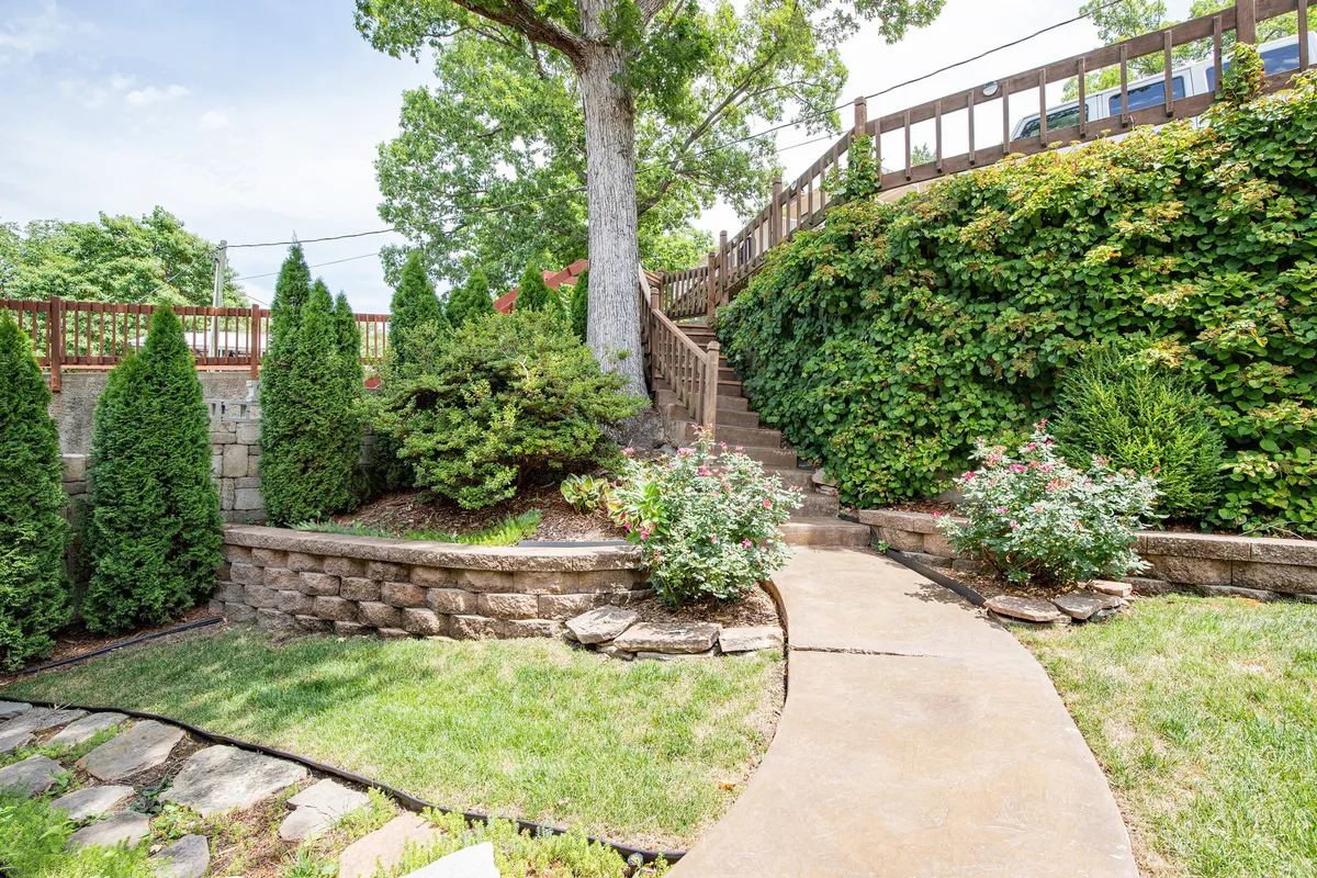 A tranquil exterior featuring a stone pathway bordered by lush greenery and flowering bushes, leading up toward the house. The scene is inviting, with dappled sunlight and hints of outdoor living—perfect for a peaceful morning coffee or an evening stroll after visiting local venues.