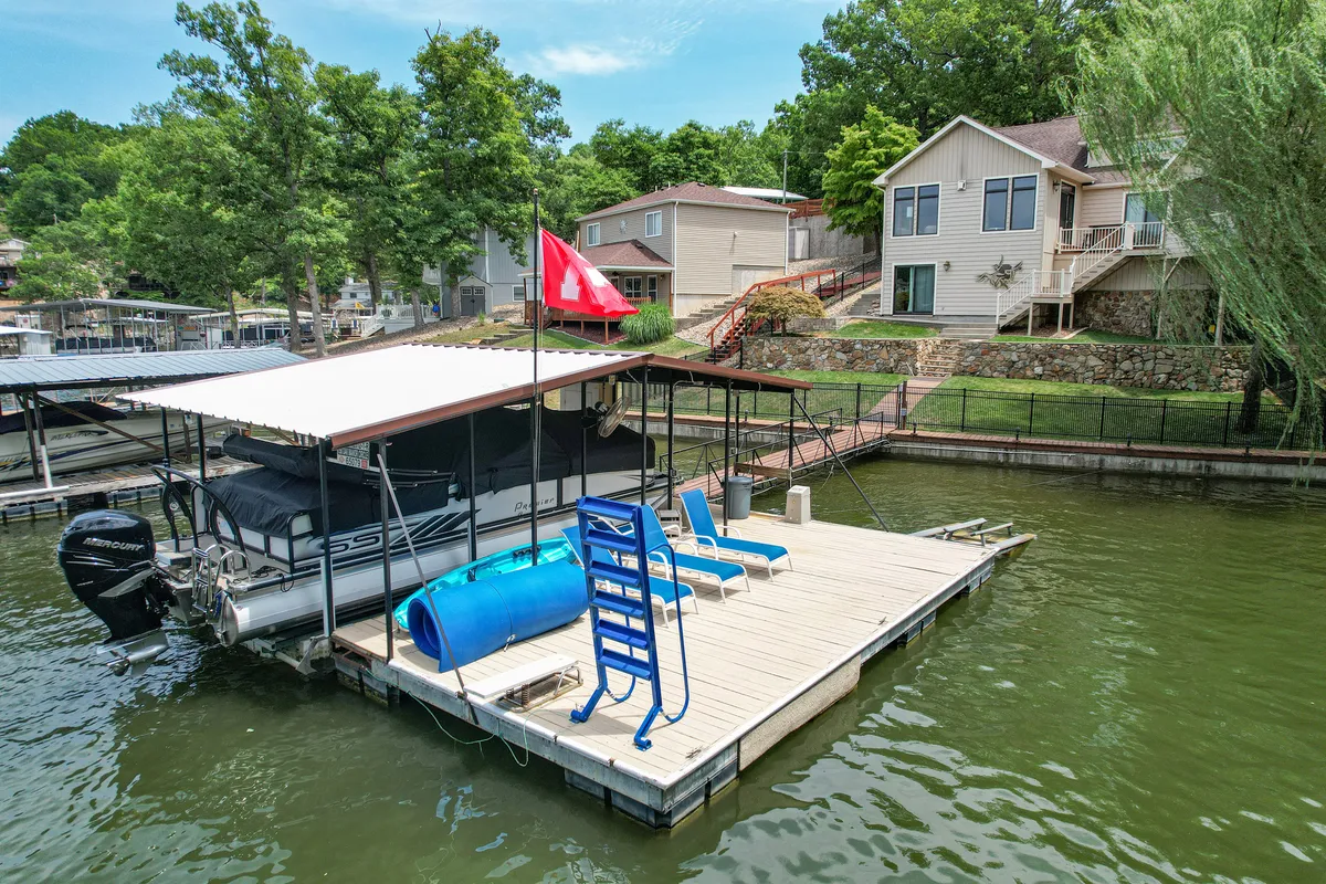 The image showcases a sun-drenched lakefront scene with a private dock extending into the sparkling water, a boat moored nearby, inviting lounge chairs perfect for relaxing, and the spacious house in the background—all framed by lush greenery.