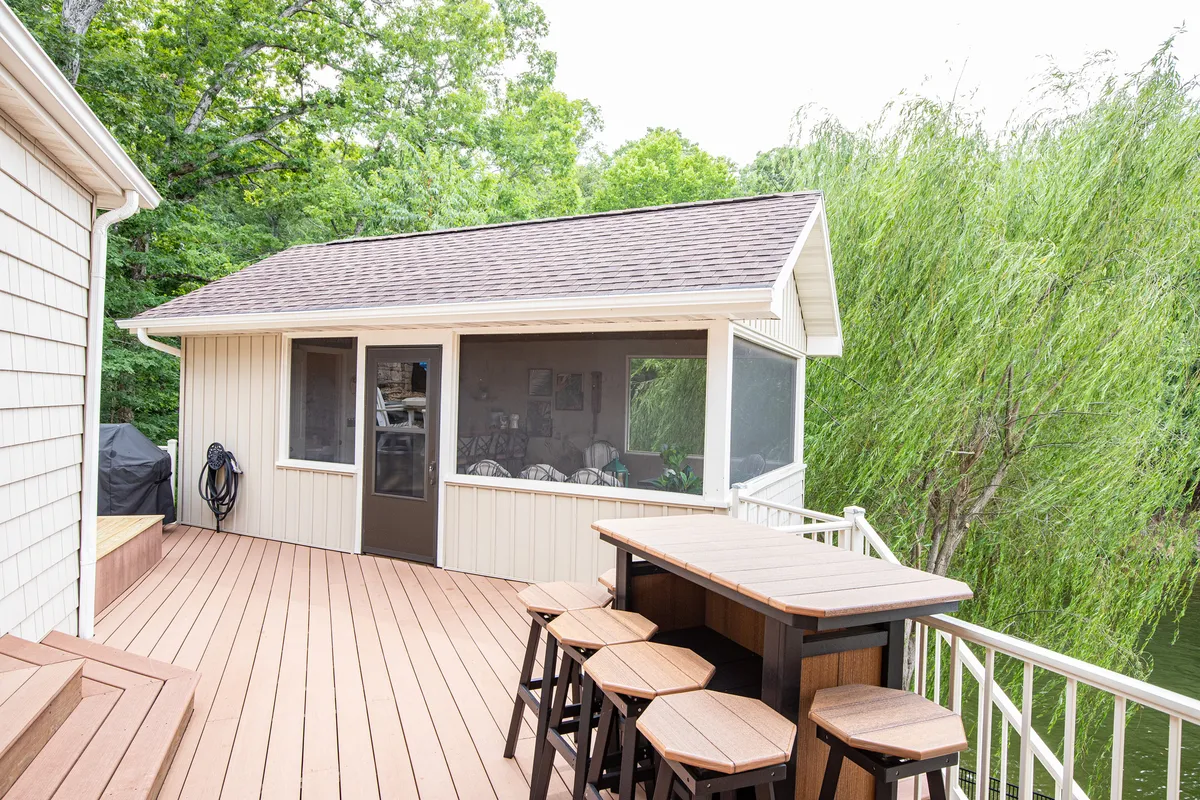 A cheerful screened-in porch featuring an outdoor bar, surrounded by lush greenery. The space is set up for social gatherings and offers a seamless connection between indoors and outdoors, with views over the lake visible through the screens.
