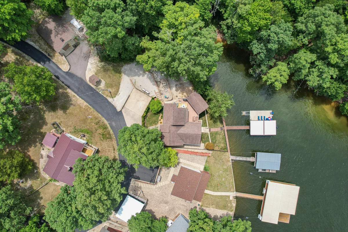 A broad exterior shot capturing the main house, the dock extending into the lake, lush surrounding greenery, and a winding road leading to the property. The setting sun casts a golden glow over the scene, hinting at a welcoming and serene environment that blends seamlessly into the Lake of the Ozarks landscape.