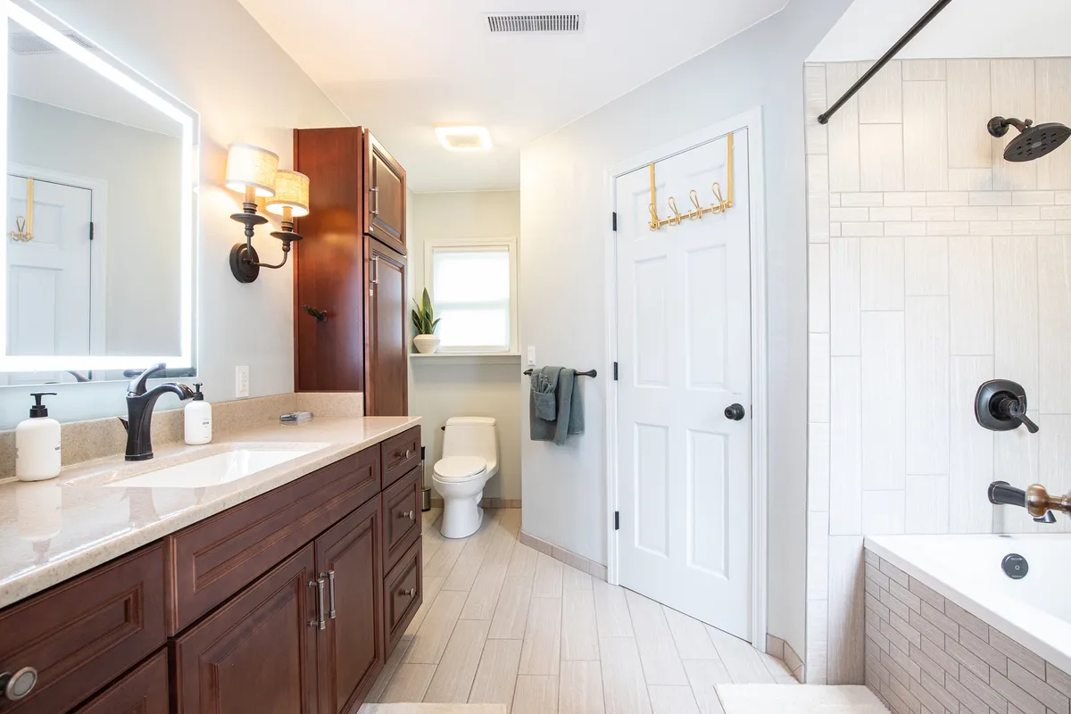A spacious, modern bathroom with a large vanity and mirror, bathtub, toilet, and a window letting in natural light. The calm color palette and spa-like ambiance evoke a sense of renewal and relaxation.