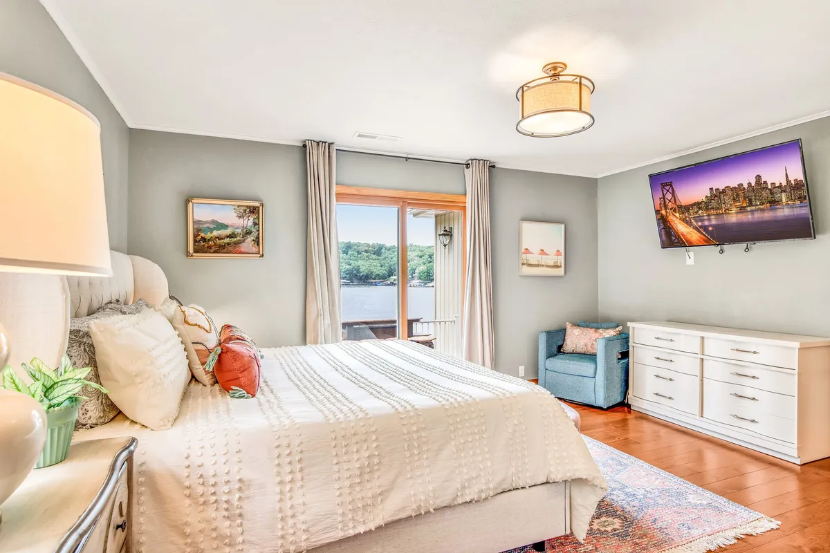 A tranquil bedroom featuring a comfortable bed positioned by a large window, a decorative lamp, and a television. The space exudes comfort with inviting textiles, soft lighting, and picturesque views outside the window.