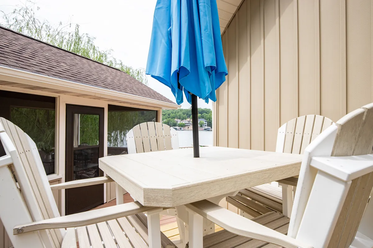The scene features a patio with a blue umbrella, a wooden table, and Adirondack chairs overlooking the water. The setting is tranquil and inviting, ideal for both casual outdoor dining and relaxed group hangouts, connecting the property’s amenities to the natural surroundings.