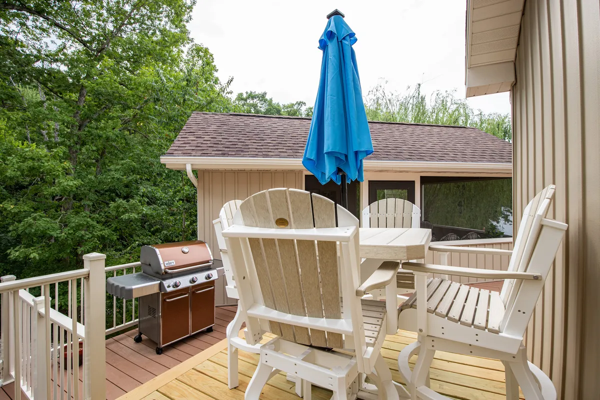 A well-appointed outdoor dining area featuring a grill, blue umbrella, and comfortable seating set on a spacious deck. The setup overlooks trees and hints at the lake nearby, evoking a sense of social gatherings, barbecues, and relaxation after exploring local restaurants or nightlife.