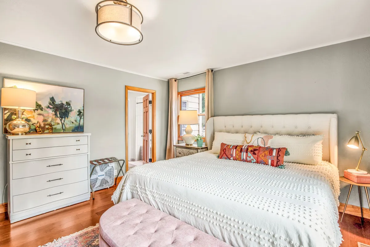 A calm, restful bedroom displaying a neatly made bed, nightstand, dresser, tasteful artwork, and a window with natural light streaming in. The subdued tones and uncluttered decor create a peaceful environment ideal for relaxation.